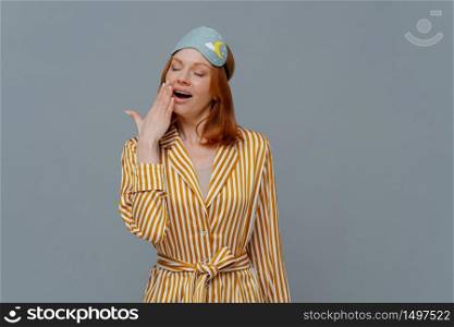 Fatigue sleepy red head woman yawns with opened mouth, wears domestic striped robe and blindfold on head, wants to sleep, stands against grey background, feels weak. Late night time concept.