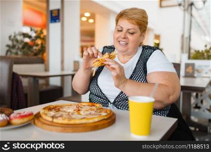 Fatty woman eating pizza in fastfood restaurant, unhealthy food. Overweight female person at the table with junk dinner, obesity problem. Fatty woman eating pizza in fastfood restaurant