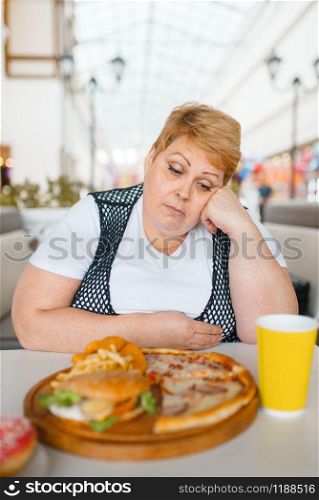 Fatty woman eating pizza in fastfood restaurant, unhealthy food. Overweight female person at the table with junk dinner, obesity problem. Fatty woman eating pizza in fastfood restaurant