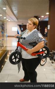 Fatty woman holding little bicycle in mall. Overweight female person with kids cycle, shopping in supermarket, obesity problem
