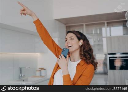 Favourite song karaoke singing at home. Happy caucasian woman in airpods is singing with mobile telephone as with microphone. Girl having fun, relaxing and dancing. Online music listening.. Favourite song karaoke singing at home. Happy caucasian woman in airpods relaxing and dancing.