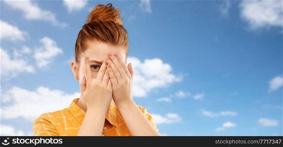 fear, vision and shame concept - scared, shy or embarrassed red haired teenage girl closing one eye by hand and looking through fingers over clouds in blue sky background. red haired teenage girl looking through fingers