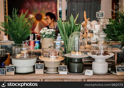 FEB 11, 2014 Chiang Mai, Thailand - Cafe interior decoration old wood counter with tropical plant pots and ceramic trays with assorted bakery pastries. Barista working behind the counter with modern wooden ceiling lamps and painting flower picture background
