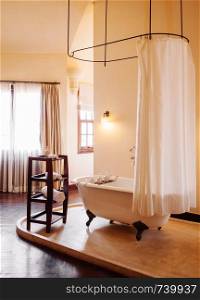 FEB 25 Dalat, Vietnam - Warm atmosphere old vintage colonial bathroom with white retro clawfoot bathtub with white curtain and wooden side table on terrazzo floor