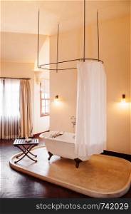 FEB 25 Dalat, Vietnam - Warm atmosphere old vintage colonial bathroom with white retro clawfoot bathtub with white curtain and wooden side table on terrazzo floor