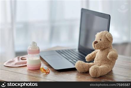 feeding and technology concept - bottle with baby milk formula, laptop computer, teddy bear toy and soother on wooden table at home. baby milk formula, laptop, soother and teddy bear