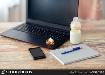 feeding and technology concept - bottle with baby milk formula, laptop computer, smartphone, notebook and soother on wooden table at home. baby milk formula, laptop and soother on table