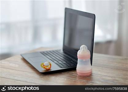 feeding and technology concept - bottle with baby milk formula, laptop computer and soother on wooden table at home. baby milk formula, laptop and soother on table