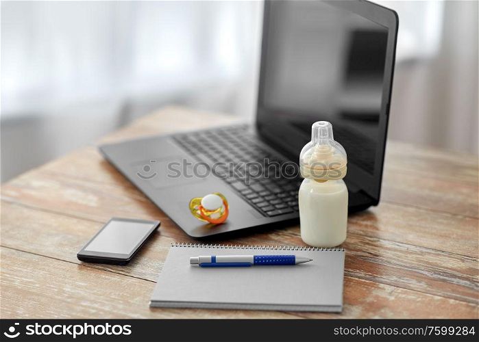 feeding and technology concept - bottle with baby milk formula, laptop computer, smartphone, notebook and soother on wooden table at home. baby milk formula, laptop and notebook on table
