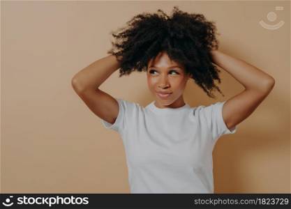 Feeling gorgeous. Portrait of young attractive sensual african woman touching with hands her curly hair and looking aside with flirty look, dressed in white basic tshirt, isolated on beige background. Young attractive sensual african woman touching with hands curly hair, isolated on beige background