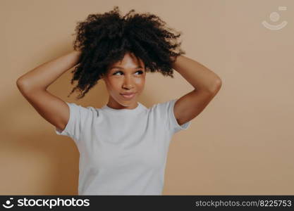 Feeling gorgeous. Portrait of young attractive sensual african woman touching with hands her curly hair and looking aside with flirty look, dressed in white basic tshirt, isolated on beige background. Young attractive sensual african woman touching with hands curly hair, isolated on beige background