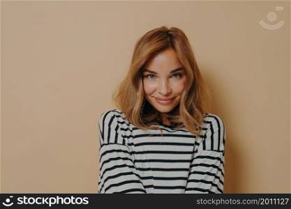 Feeling shy. Young charming woman with dyed blonde hair in striped shirt playfully smiling while looking at camera with head slightly down, isolated over beige studio background with free space. Young charming woman with dyed blonde hair in striped shirt looking at camera with shy smile