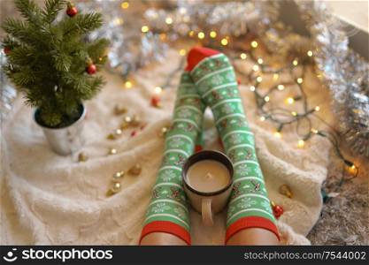 Feet in christmas socks near the Christmas tree. Woman sitting at the blanket, drinks hot beverage and relaxes warming up their feet in woollen socks. Winter and Christmas holidays concept