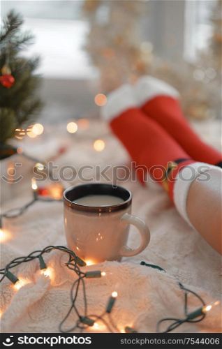 Feet in Santa&rsquo;s socks near the Christmas tree. Woman sitting at the blanket, drinks hot beverage and relaxes warming up their feet in woollen socks. Winter and Christmas holidays concept