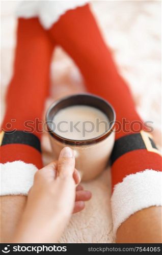 Feet in Santa&rsquo;s socks near the Christmas tree. Woman sitting at the blanket, drinks hot beverage and relaxes warming up their feet in woollen socks. Winter and Christmas holidays concept