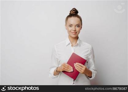 Female accountant in white shirt with hair in bun holding red notebook that contains logs and transactions of her company and looking at camera with confidence, standing isolated on grey background. Female accountant in white shirt and notebook