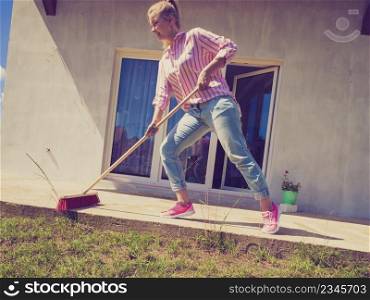 Female adult young woman using big broom to clean up backyard patio. Housecare duties concept.. Woman cleaning patio using brush broom