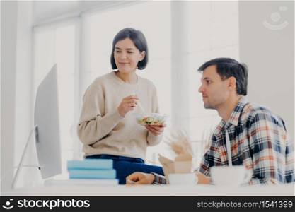 Female and male colleagues collaborate together, eat salad, have lunch time, concentrated into computer, pose in coworking space, search necessary information and prepare project for meeting