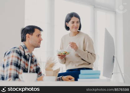 Female and male colleagues collaborate together, eat salad, have lunch time, concentrated into computer, pose in coworking space, search necessary information and prepare project for meeting