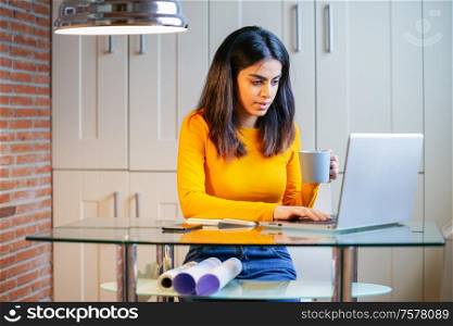 Female architect working at home with a laptop and blueprints. Persian woman. Persian woman with a coffee mug.. Female architect working at home with a laptop and blueprints
