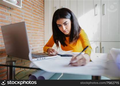 Female architect working at home with a laptop and blueprints. Persian woman designing a project.. Female architect working at home with a laptop and blueprints