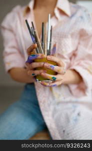 Female artist washing paintbrush equipment in glass of water. Selective focus on painter hands holding drawing supply. Spare time and art creativity concept. Female artist washing paintbrush in glass of water selective focus