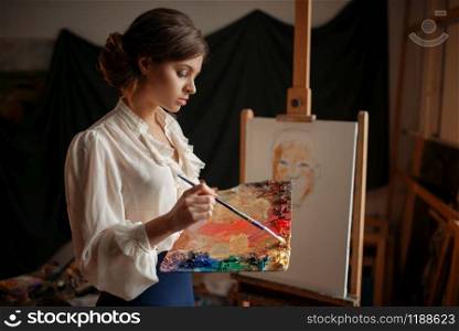 Female artist with color palette and brush standing against easel in studio. Creative paint, painter drawing portrait, workshop interior on background