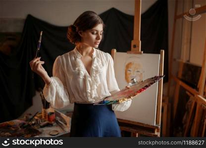 Female artist with color palette and brush standing against easel in studio. Creative paint, painter drawing portrait, workshop interior. Female artist with color palette and brush