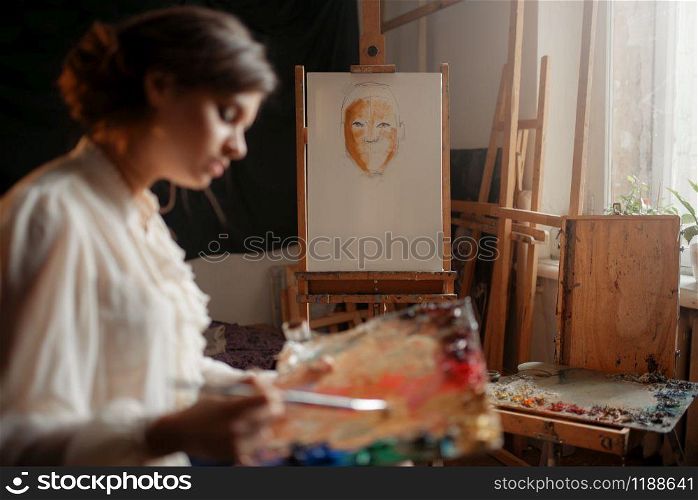 Female artist with color palette and brush standing against easel in studio. Creative paint, painter drawing portrait, workshop interior. Female artist with color palette and brush