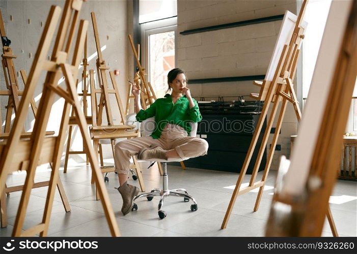 Female artist with paintbrush in hand looking seriously on her own picture while sitting on chair in art studio. Female artist with paintbrush in hand looking seriously on her own picture