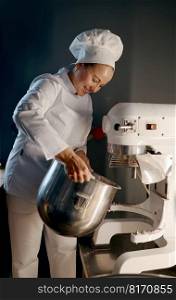 Female baker wearing uniform working in professional kitchen with large dough mixer at bakery. Female baker working in professional kitchen with dough mixer at bakery