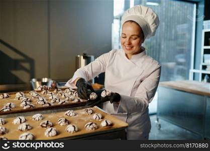 Female baker wearing white uniform laying out freshly cooked cookies. Baking tray with sweet pastry. Female baker in uniform laying out freshly cooked cookies