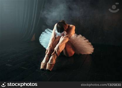 Female ballet performer in white dress sits on the floor, body flexibility. Ballerina training in dancing class