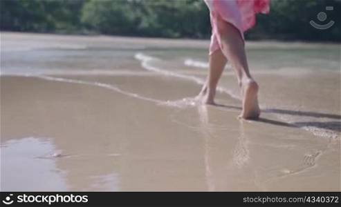 female barefoot walking along the island beach with clear sea water waves in slow motion, Splashing of water hitting legs. rear view low camera angle, summer travel booking, tropical climates country