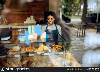 Female barista in apron at the showcase with desserts in cafe. Woman choosing sweets in cafeteria, waiter at the counter in bar. Female barista at showcase with desserts in cafe