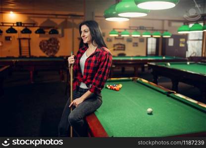 Female billiard player with cue poses at the table with colorful balls. Woman plays american pool game in sport bar. Female billiard player with cue poses at the table