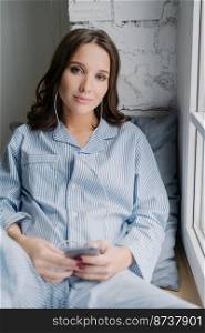 Female blogger in domestic clothes listens music in earphones and chats via cellular while sits on window sill, has calm look expression, looks seriously directly at camera, poses at home or apartment