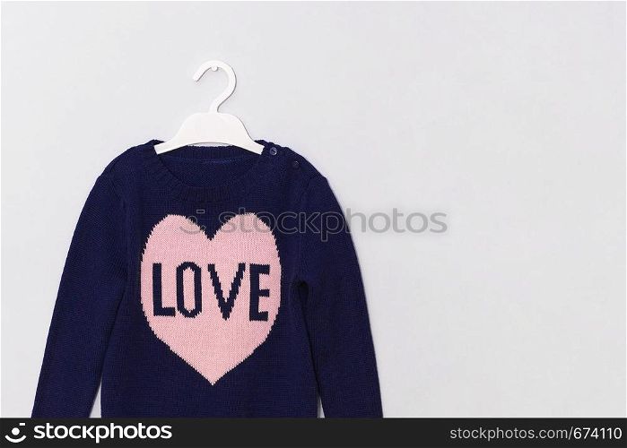 Female blue woolen sweater with pattern pink heart and inscription Love on hanger on gray background. Woman fashion look clothes concept. Copy space.. Female blue woolen sweater with pattern pink heart and inscription Love on hanger on gray background. Woman fashion look clothes concept. Copy space