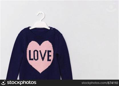 Female blue woolen sweater with pattern pink heart and inscription Love on hanger on gray background. Woman fashion look clothes concept. Copy space.. Female blue woolen sweater with pattern pink heart and inscription Love on hanger on gray background. Woman fashion look clothes concept. Copy space