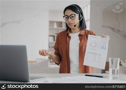 Female business coach in glasses, headset conducts online lesson by video call on laptop, showing infographic. Businesswoman holding document with charts discusses work project remotely. E-learning.. Female business coach in headset conducts online lesson by video call on laptop, showing infographic