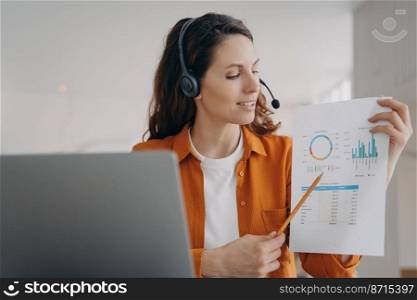 Female business coach in headset with microphone conducts video call negotiations with partners at laptop. Businesswoman employee showing chart, presenting project online on working conference.. Female employee in headset showing chart on online working conference, conducts video call at laptop