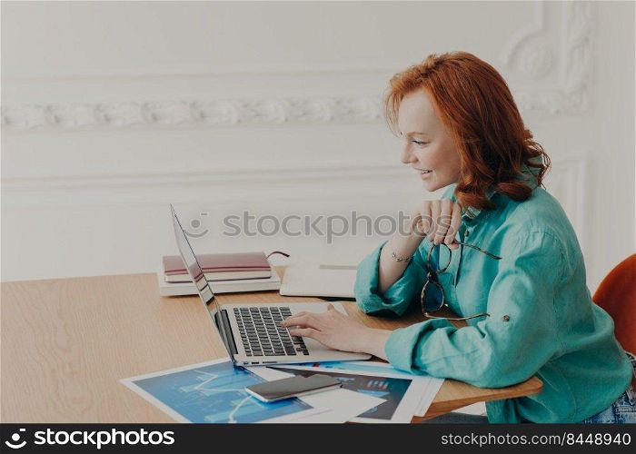 Female business owner prepares startup project, browses internet on laptop, works freelance, poses in workplace at home, does distant job, chats with colleagues has pleased expression dressed casually