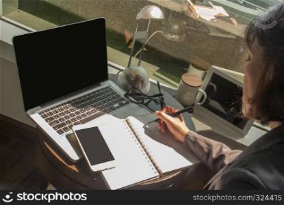 Female businessmen are taking notes in a notebook while watching a job on a laptop with glasses, lamps, coffee cups and tablets and placed on the table.