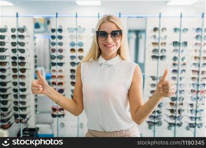 Female buyer tries on sunglasses in optics store, showcase with spectacles on background. Eyes protection from sunlight in glasses shop, eyecare concept