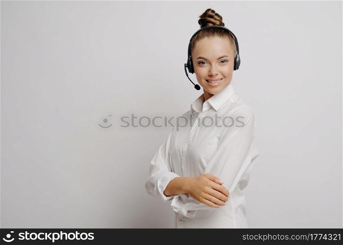 Female call center agent in classic wear, woman in black headset connecting with customer, standing sideways on light backgroun and looking with confidence at camera, copy space for text. Female call center agent with black headset and crossed arms