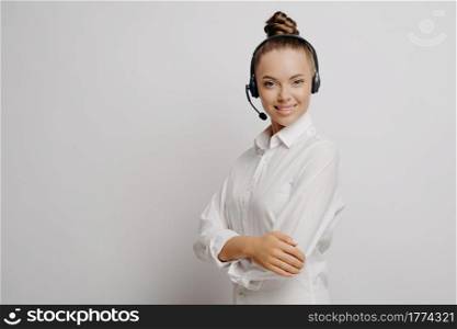 Female call center agent in classic wear, woman in black headset connecting with customer, standing sideways on light backgroun and looking with confidence at camera, copy space for text. Female call center agent with black headset and crossed arms