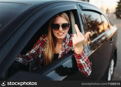 Female car driver in sunglasses shows an obscene gesture. Impudent woman in vehicle, aggressive automobile driving concept. Female car driver shows an obscene gesture