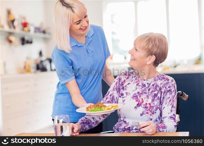 Female Care Assistant Serving Meal To Senior Woman Seated In Wheelchair At Table