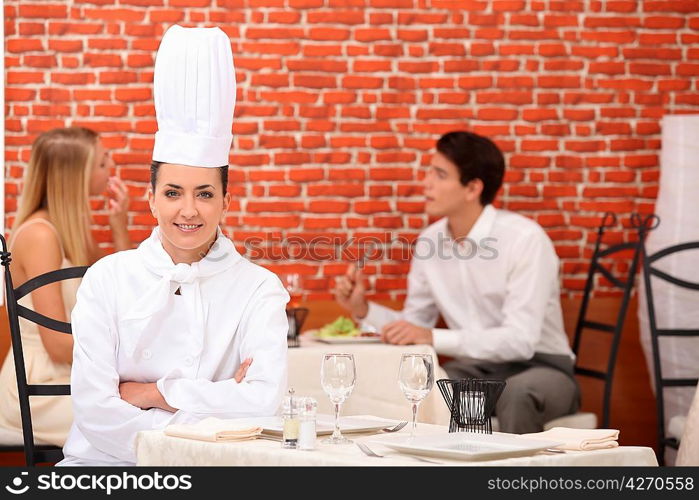 female chef posing in restaurant with couple dining in background