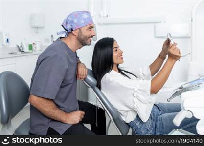 Female client looking at his teeth in the mirror next to a dentist in a room of a dental clinic. Client looking at his teeth in the mirror next to a dentist in a clinic